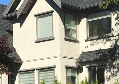 Close-up of a beige stucco home with grey-framed windows and blinds