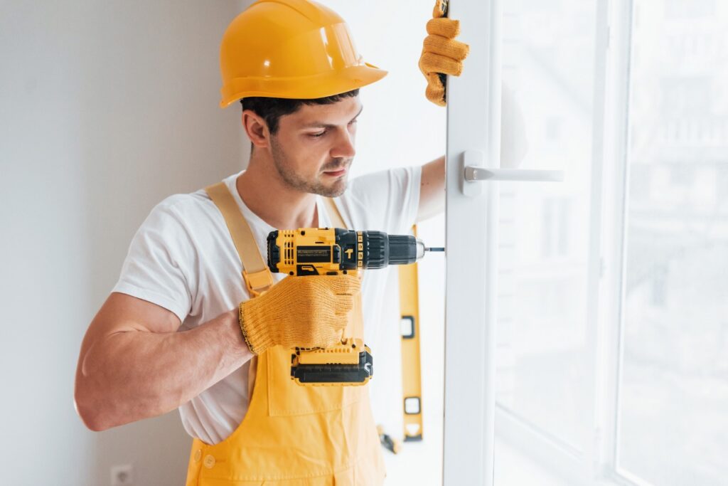 Installer in a hard hat holding a drill and level beside a window frame